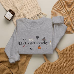 Puede incluir: Una sudadera gris jaspeado de cuello redondo con el texto "Let's get spooky!" en negro, rodeada de pequeños iconos de Halloween que incluyen una lápida, murciélagos, huesos, un fantasma, un sombrero de bruja, una vela, piruletas, una calabaza y una araña.