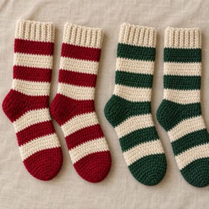 Può includere: Quattro calzini all'uncinetto disposti in fila. Due calzini presentano strisce orizzontali rosso scuro e crema, mentre gli altri due presentano strisce orizzontali verde bosco scuro e crema. Ogni calzino ha un bordo a coste sull'apertura superiore.