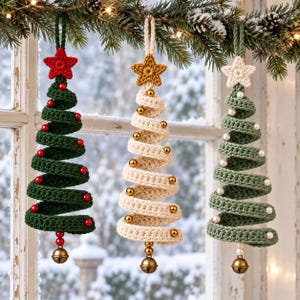 Könnte beinhalten: Drei spiralförmige Häkel-Weihnachtsbaumschmuckstücke, die an einem Tannenzweig hängen. Der linke Baum ist dunkelgrün mit roten Perlen und einem roten Stern. Der mittlere Baum ist cremefarben mit goldenen Perlen und einem goldenen Stern. Der rechte Baum ist salbeigrün mit weißen Perlen und einem weißen Stern. Jeder Baum hat eine Metallglocke an der Basis.