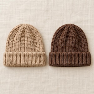 Könnte beinhalten: Zwei Strickmützen, eine in hellem Beige und eine in Dunkelbraun. Beide Mützen weisen eine gerippte Textur über die gesamte Krone auf und verfügen über einen breiten, umgeschlagenen Saum am unteren Rand mit einem durchgehenden vertikalen Strickmuster.