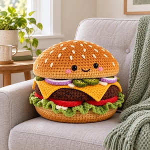 Könnte beinhalten: Ein rundes, gehäkeltes Hamburger-Plüschtier mit einem beigen Brötchen mit weißen gestickten Sesamsamen, einem braunen Patty, gelbem Käse, roten Tomatenscheiben, violetten Zwiebelringen, grünen Gewürzgurken und einer Basis aus krausem grünem Salat.