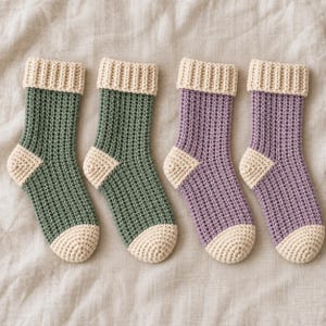 Könnte beinhalten: Vier gehäkelte Socken in einer Reihe. Zwei Socken sind dunkelgrün mit cremefarbenen Bündchen, Fersen und Zehenspitzen. Die anderen beiden Socken sind hellviolett mit cremefarbenen Bündchen, Fersen und Zehenspitzen. Jede Socke hat ein gitterartiges Muster.
