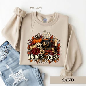 May include: A sand-colored crewneck sweatshirt featuring a graphic print of a skull, pumpkins, a steaming mug with a crescent moon, books, and autumn leaves. The text "UNHOLY AND COZY" is printed below the illustration in a decorative font.