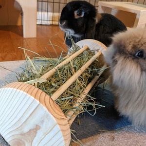 May include: A wooden hay feeder filled with hay, with two rabbits eating from it. The feeder is made of light-colored wood and has a cylindrical shape with wooden bars. One rabbit is black and the other is brown.