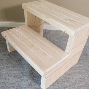 May include: A two-step wooden step stool made of unfinished pine. The steps have grooved surfaces for grip. The stool is designed with a simple, sturdy construction, ideal for reaching higher places. The natural wood grain is visible throughout.