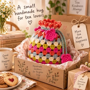 Puede incluir: Una bolsa de ganchillo multicolor con cordón y un detalle de flor, dentro de una caja de regalo de cartón. La caja tiene el texto 'Handmade Goodness for Brighter Days' y una etiqueta que dice 'More tea More happiness'. Una taza y galletas cerca.