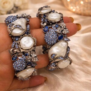 May include: Two ornate bangles featuring large, irregular white pearls set in a dark metal frame. The surface is densely encrusted with small, light blue and dark blue crystals, along with clusters of tiny white seed pearls throughout the design.