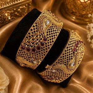Peut inclure: Deux bracelets rigides en métal doré ornés de motifs complexes en filigrane et de paons en relief. Le bijou est agrémenté de petites pierres rouges en forme de goutte et de cristaux blancs étincelants incrustés dans la surface texturée.
