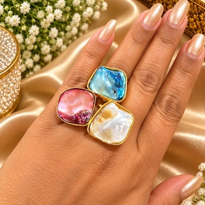 May include: A gold-toned ring featuring three irregular, square-shaped cabochons made of iridescent shell material. The stones are arranged in a cluster, displaying vibrant shades of blue, pink, and white with a polished, reflective finish.
