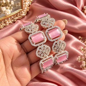 Puede incluir: Un par de brazaletes plateados con piedras rosas rectangulares alternadas con motivos florales circulares incrustados con pequeños cristales transparentes. Las piezas de joyería tienen un marco metálico y un patrón repetitivo.