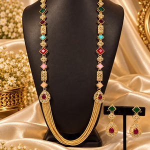 May include: A gold-toned necklace and earring set featuring multi-colored geometric stones including green, red, blue, and pink accents. The long necklace has a multi-strand chain base with a teardrop pendant, while the matching earrings feature a square green stone top and a teardrop-shaped red stone drop.