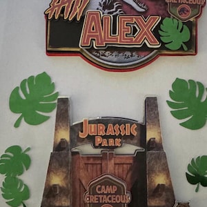 May include: A decorative set featuring a Jurassic Park gate cutout with the text Camp Cretaceous, a circular sign with a dinosaur silhouette and the name Alex, and several green paper monstera leaves arranged around the central elements.