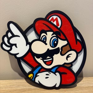 May include: A circular wall sign featuring a colourful graphic of the character Mario. The design includes a red hat with a white M, blue overalls, a black moustache, and white gloves. Mario is pointing upwards with one hand against a white background.