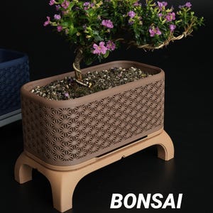May include: A bonsai tree with small purple flowers planted in a rectangular brown planter featuring a geometric lattice pattern. The planter rests on a light tan stand with four legs. The text BONSAI Kumiko Collection is visible below.