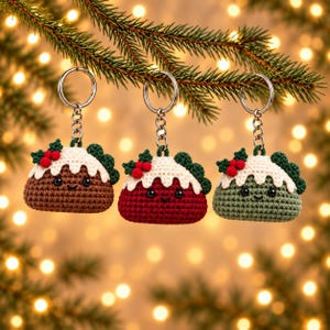 May include: Three crochet keychains shaped like Christmas puddings in brown, red, and green. Each features a white icing top, dark green holly leaves, three red berries, and black bead eyes. The keychains are attached to silver metal rings.