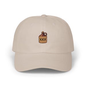 May include: A beige baseball cap featuring an embroidered graphic of a brown ceramic jug with a handle and a cork stopper. The jug has the letters XXX stitched in black on its front surface. The cap has a curved brim and a structured crown.