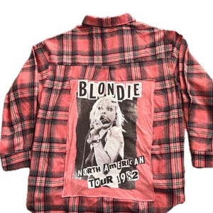 May include: A long-sleeve button-down shirt in a red and black plaid pattern. The back features a large rectangular patch with a black and white photograph of a singer holding a microphone, with the text BLONDIE NORTH AMERICAN TOUR 1982.