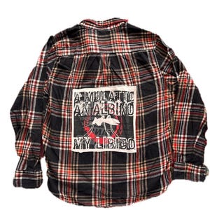 May include: A long-sleeve button-down shirt featuring a black, red, and white plaid pattern. A large white rectangular patch is sewn onto the back, displaying a black graphic of a mosquito inside a red circle with the text A MULATTO AN ALBINO MY LIBIDO.