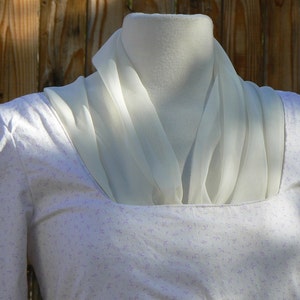 May include: A light-colored dress with a square neckline and long sleeves, adorned with a delicate floral pattern. A sheer, cream-colored scarf is draped around the neck, adding a touch of elegance to the garment. The dress is displayed on a mannequin.