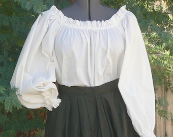 May include: A white off-the-shoulder blouse with long, gathered sleeves and a dark green skirt. The blouse has a ruffled neckline and cuffs. The skirt is pleated. The outfit is displayed on a mannequin.