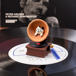 May include: A miniature brown gramophone figurine featuring a white cat with a black ear patch inside the horn. The device has a black record on its base and a small brown crank handle on the side, resting on a large black vinyl record.