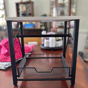 May include: A small, rectangular storage stand featuring a white top surface supported by a black metal frame. The unit includes a lower shelf made of black wire mesh with a cutout front panel. The text GrainWoodDecor is visible.