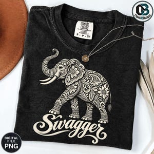 May include: A black short-sleeve t-shirt featuring a graphic of an elephant decorated with intricate floral and swirl patterns. Below the elephant, the word "Swagger" is printed in a white cursive script. The shirt has a Comfort Colors label.