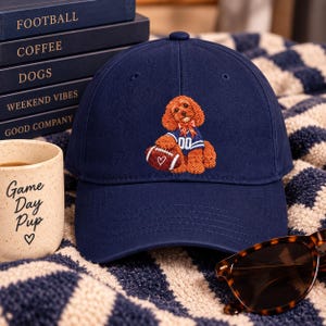 May include: A navy blue baseball cap featuring an embroidered design of a brown curly-haired dog wearing a blue jersey with the number 00 and a red bandana, holding a brown football with a white heart graphic on the front of the crown.