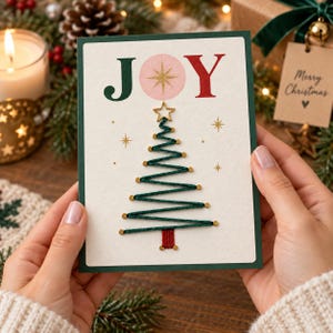 Puede incluir: Una tarjeta de felicitación blanca con borde verde oscuro que presenta el texto JOY en letras verdes y rojas. La letra O se sustituye por un círculo rosa con una estrella dorada. Un árbol de Navidad estilizado formado por hilo verde y tachuelas.