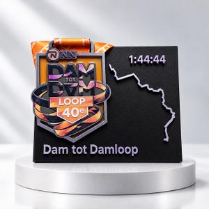 Könnte beinhalten: Eine schwarze rechteckige Präsentationsplatte mit einer metallischen Dam tot Damloop Medaille und einem orangefarbenen Band. Die Platte zeigt den Text 1:44:44, eine weiße Linienkartengrafik und den Text Dam tot Damloop in helllila Schrift unten.
