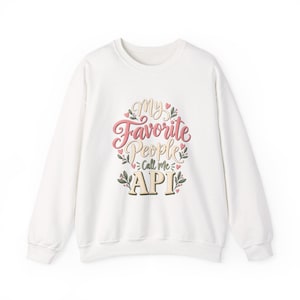 May include: A white long-sleeve crewneck sweatshirt featuring a central graphic design with the text My Favorite People Call Me API in pink, beige, and dark green lettering, surrounded by small pink hearts and green leaf illustrations.