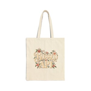 May include: A cream-colored canvas tote bag featuring the text "BLESSED API" in a stylized, serif font. The lettering is surrounded by a decorative illustration of small pink flowers, green leaves, and thin brown stems centered on the front.