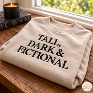 Puede incluir: Una sudadera beige de cuello redondo con el texto bordado en negro "TALL, DARK & FICTIONAL" en el pecho. La prenda tiene un cuello acanalado y mangas largas, presentada en una posición plegada sobre una superficie de madera.