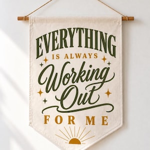 May include: A cream-colored fabric wall banner suspended from a wooden dowel with a rope. The banner features the dark green and mustard yellow text "EVERYTHING IS ALWAYS Working Out FOR ME" with small star accents and a sun icon at the bottom.