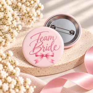 Könnte beinhalten: Ein runder Ansteckbutton mit hellrosa Hintergrund und dem Schriftzug Team Bride in dunkelrosa Schreibschrift. Unter dem Text befindet sich eine Illustration einer rosa Schleife. Die Rückseite ist aus silbernem Metall mit einer Nadel.