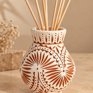 May include: A terracotta reed diffuser vessel featuring a white matte finish with intricate carved patterns revealing the natural reddish-brown clay underneath. The bulbous container has a flared rim and holds several thin wooden reeds.