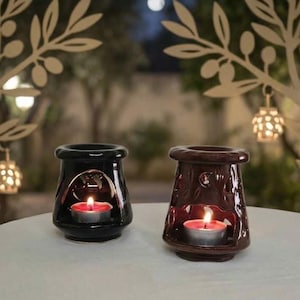 May include: Two ceramic oil burners, one black and one dark brown, each featuring a wide opening at the base for a tea light candle. The burners have a smooth, glossy finish and a flared top section designed to hold oil or wax melts.