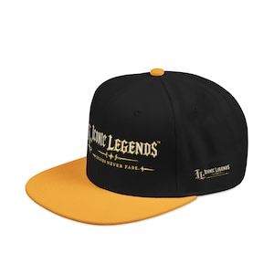 Könnte beinhalten: Eine schwarze Snapback-Cap mit einem leuchtend gelben Schirm und einem passenden gelben Knopf an der Oberseite. Auf der Vorderseite befindet sich der goldene Schriftzug Iconic Legends und der Satz Legends Never Fade. Seitlich ist ein kleineres Logo.