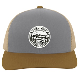 May include: A grey and tan trucker hat featuring a circular patch on the front. The patch displays a black and white graphic of the Great Smoky Mountains National Park, including mountain peaks, pine trees, a winding road, and stars.