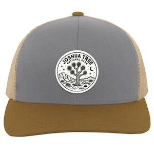 May include: A grey and tan trucker hat featuring a circular white patch on the front. The patch displays a black line art illustration of a Joshua tree, desert landscape, stars, and a crescent moon with the text Joshua Tree National Park.