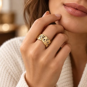 Puede incluir: Un anillo de banda ancha en color dorado con una superficie irregular y texturizada que presenta detalles negros en relieve. El anillo tiene un perfil de borde ondulado y orgánico con un acabado mate, destacando el contraste entre el oro y los tonos oscuros.