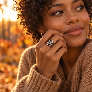 Puede incluir: Un anillo de plata con una piedra de color gris oscuro de forma orgánica e irregular, montada en una banda de metal pulido. El anillo presenta un engaste texturizado e irregular que enmarca la piedra facetada, creando un contraste entre el acabado metálico y el centro oscuro.