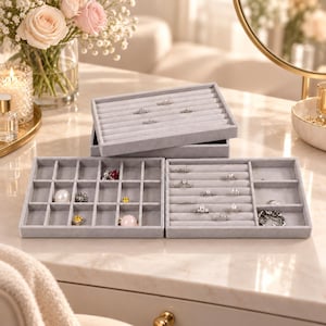 May include: A set of three light grey velvet jewellery organiser trays. One tray features multiple long slots for rings, whilst the other two trays are divided into various square and rectangular compartments, containing assorted rings and small jewellery pieces.