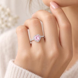 Puede incluir: Un anillo de tono plateado con una piedra preciosa central de color rosa claro en forma de corazón, rodeada por un halo de pequeñas piedras transparentes. La banda es fina y está incrustada con pequeñas piedras transparentes visibles.
