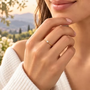 Pode incluir: Um anel fino de ouro com uma pequena pedra preciosa redonda e transparente cravada no centro. A banda é lisa e polida, apresentando um design simples que destaca a pedra solitária. O anel é usado num dedo, exibindo o seu perfil delicado.