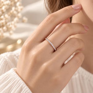 Könnte beinhalten: Ein schmaler Ring aus Roségold mit einer durchgehenden Reihe kleiner, runder, klarer weißer Steine, die entlang des gesamten äußeren Umfangs gefasst sind. Das Metall hat einen warmen, metallisch rosafarbenen Ton und die Steine sind eingefasst.
