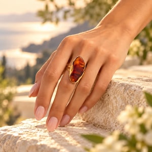 Puede incluir: Un anillo de oro con una piedra de ámbar grande, irregular y pulida en tonos naranja y marrón intenso. El engaste tiene un acabado metálico texturizado y orgánico que sigue la forma natural y asimétrica de la piedra de ámbar.