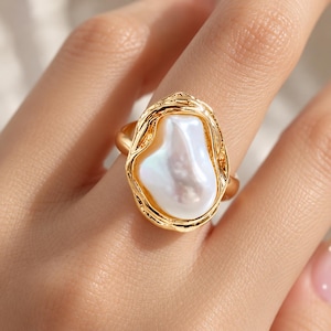 Puede incluir: Un anillo de tono dorado con una perla barroca blanca grande e irregular, engastada en un marco de metal texturizado de forma orgánica. La banda es lisa y pulida, contrastando con los detalles intrincados e irregulares que rodean la perla.