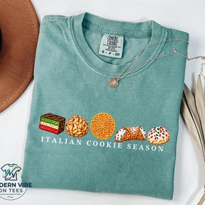 May include: A sage green t-shirt featuring a graphic of five assorted Italian cookies arranged in a row above the white text "ITALIAN COOKIE SEASON". The shirt has a crew neckline and a Comfort Colors brand label printed on the inside collar.