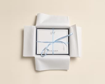 May include: A stack of white stationery cards with a dark border, a monogram at the top, and the text RACHEL & JACK at the bottom, tied with a light blue ribbon and nestled inside an open white paper gift box.